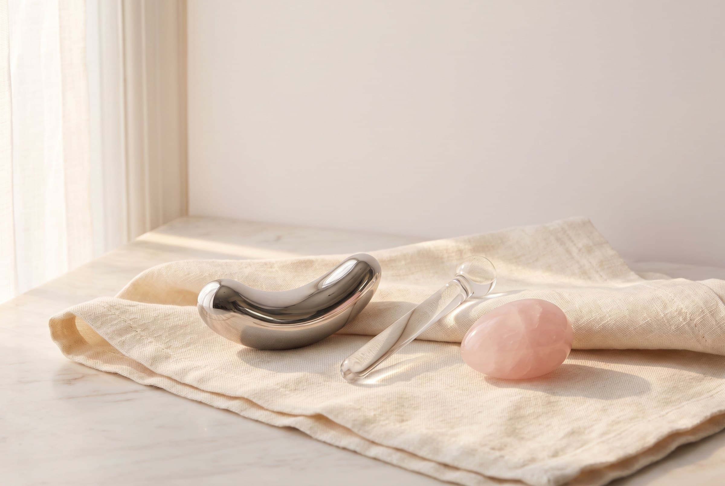 A polished stainless steel form, a clear glass wand and a smooth rose-quartz egg resting on cream linen in soft morning light