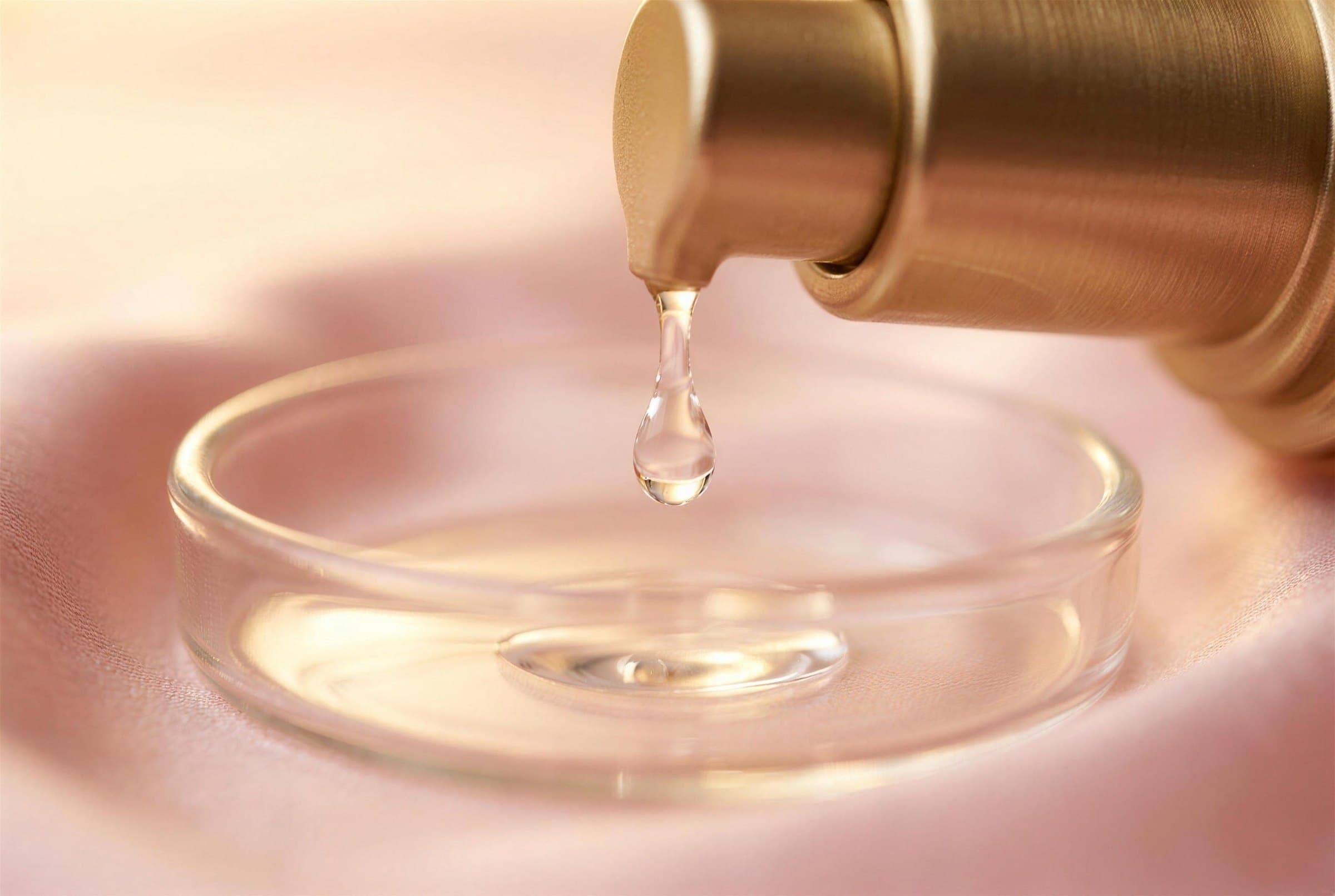 A clear drop of lubricant falling from a brushed gold pump into a small glass dish on blush silk, lit by warm golden light